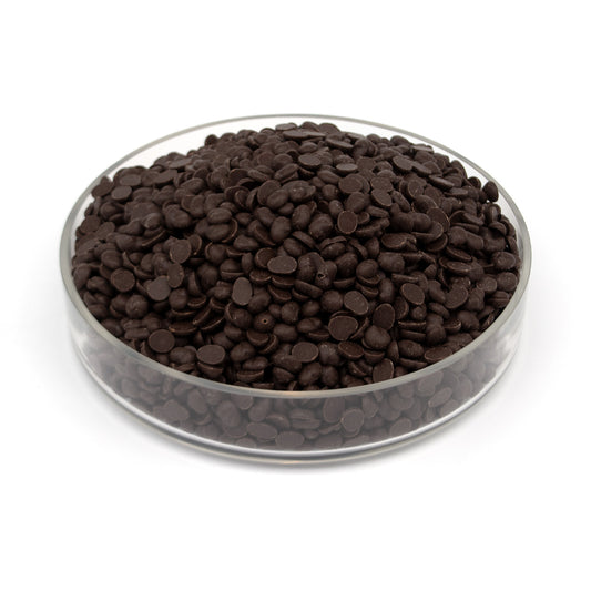 A pile of small dark chocolate discs in a clear glass bowl on a white background.