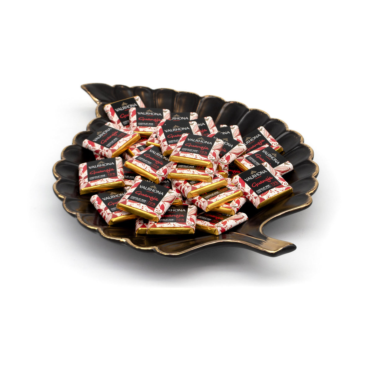 A decorative leaf-shaped black dish with gold-toned edges, filled with individually wrapped Valrhona Guanaja chocolate squaress on a clean white background.