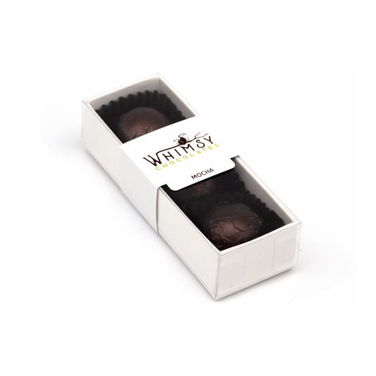A white rectangular chocolate box with a clear lid, revealing mocha chocolates inside dark paper cups. The lid has a white label with the brand name 'Whimsy Chocolates' and the word 'MOCHA' printed on it, set against a plain white background.